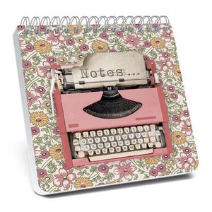 May include: A spiral-bound notepad with a floral pattern cover. The notepad features a pink typewriter illustration with the word "Notes..." printed on a paper sheet. The keyboard keys are white.