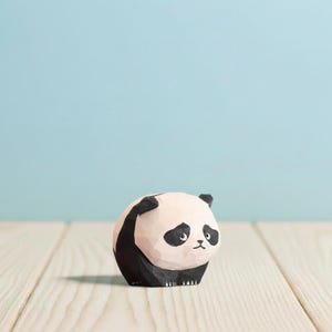 May include: A wooden panda figurine with a light pink body, black accents, and a detailed face. The panda is sitting on a light wood surface against a light blue background. The panda's black markings include its ears, eyes, and parts of its body.