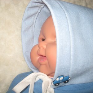 May include: A baby doll wearing a light blue hooded outfit with a white tie and a blue truck applique. The doll has a rosy complexion and is wearing a white hat.