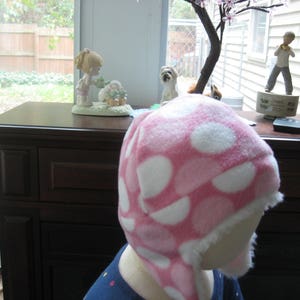 May include: A pink and white polka dot fleece hat with a white fur trim. The hat has ear flaps and a pom pom on top.