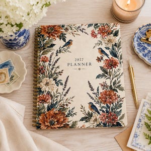 May include: A spiral-bound 2027 planner with a cream-colored cover featuring a botanical illustration of rust-colored flowers, green foliage, and small blue birds. The text 2027 PLANNER is printed in the center in a dark, elegant serif font.