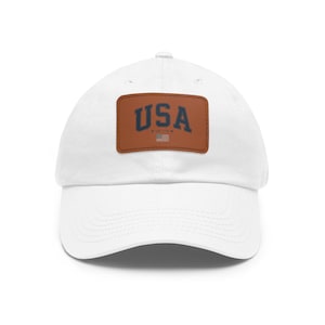 Pode incluir: Boné de beisebol branco com aba curva. Um patch de couro marrom na frente exibe a palavra "USA" em azul marinho, com "EST. 1776" e uma pequena bandeira americana. Um acessório casual.