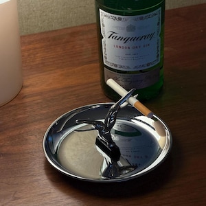 May include: A circular, polished silver-tone metal ashtray featuring a central sculptural figure of a diver in a mid-air pose. The figure serves as a rest for a single cigarette. The ashtray has a reflective surface and a smooth, rounded rim.