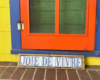 May include: A wooden sign with blue tile letters spelling "JOIE DE VIVRE" on a brick patio in front of a red door with a green screen door. The door is framed by blue and yellow painted trim.