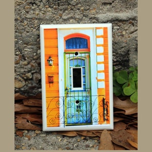 May include: A light switch cover with a colorful image of a door with a blue and green design. The door has a wrought iron gate and a small window. The background is a yellow and white brick wall.