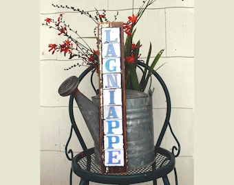 May include: A wooden sign with the word "LAGNIAPPE" spelled out in blue and white tiles. The sign is sitting on a chair with a watering can and flowers.