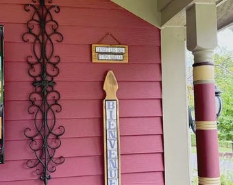 May include: A tall black wrought iron wall scroll, a rectangular wooden sign with the text "LAISSEZ LES BON TEMPS ROULER", and a vertical wooden picket sign with the word "BIENVENUE" in blue letters, all mounted on a red horizontal siding wall.