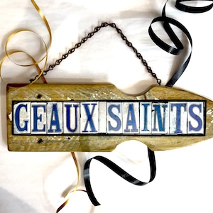 May include: A wooden sign with the words "GEAUX SAINTS" spelled out in blue and white tiles. The sign is hanging from a chain and has a rustic, weathered look.