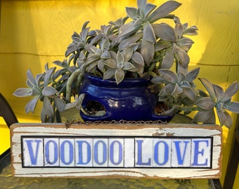 May include: A wooden sign with the words "Voodoo Love" in blue letters on white tiles. The sign is hanging from a chain and is in front of a blue pot with a succulent plant.