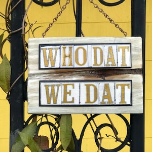 May include: A white wooden sign with black trim hangs from a chain. The sign has two lines of gold letters that read "WHO DAT" and "WE DAT".