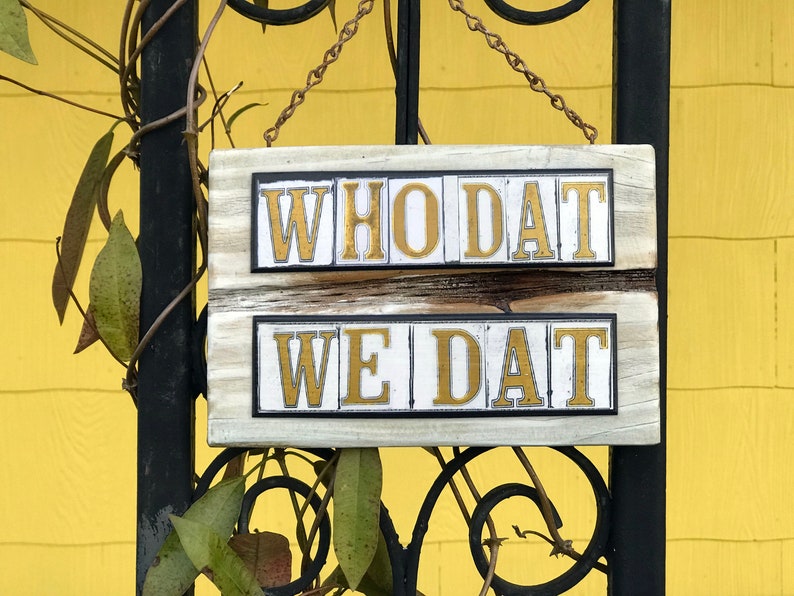 May include: A white wooden sign with black trim hangs from a chain. The sign has two lines of gold letters that read "WHO DAT" and "WE DAT".