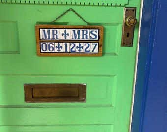 May include: A rectangular wooden sign hanging by a chain, featuring two rows of white tiles with blue text and fleur-de-lis symbols. The top row reads MR and MRS, while the bottom row displays the date 06-12-27 in a blue font.
