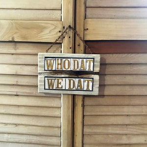 May include: A white wooden sign with black lettering hangs on a wooden door. The sign reads "WHO DAT WE DAT".