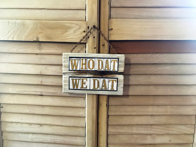 May include: A white wooden sign with black lettering hangs on a wooden door. The sign reads "WHO DAT WE DAT".