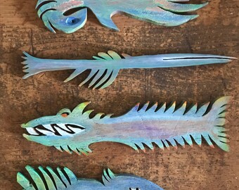 May include: Four wooden fish ornaments painted in shades of blue, green, and purple. The fish have different shapes and sizes, with some having teeth and fins.