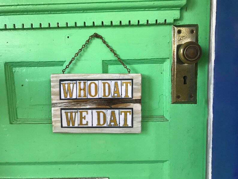 May include: A wooden sign with the words "WHO DAT WE DAT" in gold letters on a white background. The sign is hanging on a green door with a brass door handle.