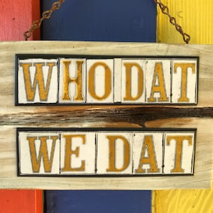 May include: A wooden sign with the words "Who Dat" and "We Dat" in gold letters on a white background.