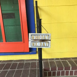 May include: A wooden sign with the words "Who Dat" and "We Dat" in gold lettering, hanging on a black metal railing in front of a red, blue, and yellow house.