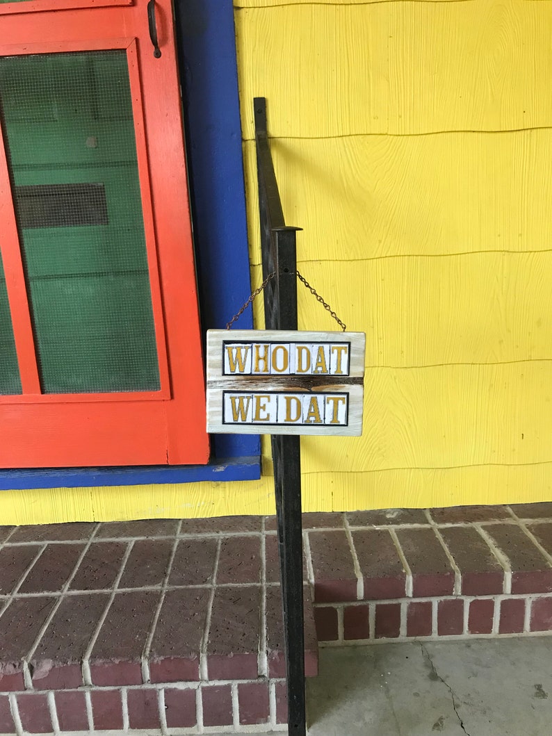 May include: A wooden sign with the words "Who Dat" and "We Dat" in gold lettering, hanging on a black metal railing in front of a red, blue, and yellow house.