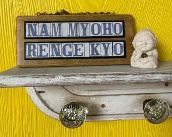 May include: A wooden sign with the text "NAM MYOHO RENGE KYO" in blue and white tiles, displayed on a distressed white shelf. A small, off-white Buddha statue sits to the right. The shelf has two glass doorknob hooks.