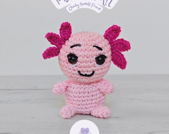 May include: A handmade, pink crocheted axolotl doll with dark pink gills and a smiling face. The doll has black eyes and a small, curved mouth. The image includes text that reads "PDF PATTERN!" and "Chunky Axolotl Friend".