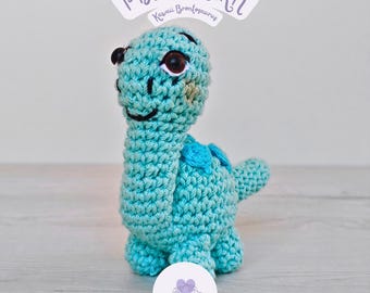 May include: A turquoise crochet dinosaur toy with brown eyes and a smiling face. The dinosaur has a long neck and a small tail. The text "PDF PATTERN" and "Kawaii Brontosaurus" are on a white banner above the dinosaur. A purple heart-shaped logo is at the bottom.