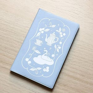 Peut inclure: Un carnet bleu clair avec un motif décoratif blanc représentant un cygne, une théière et des éléments floraux. Le carnet est rectangulaire et fermé, posé sur une surface en bois.