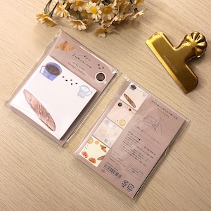 Peut inclure: Deux blocs-notes autocollants avec des illustrations d'aliments, dont un croissant, une tasse de café et une baguette. Un clip doré et un bouquet de fleurs jaunes sont également présents.