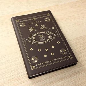 Peut inclure: Un carnet marron avec des détails dorés, affichant le mot "coffee" en anglais et en japonais. La couverture présente une illustration de moulin à café et des motifs de grains de café. Le carnet est fermé et posé sur une surface en bois clair.
