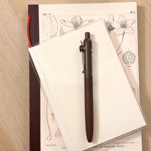Może przedstawiać: Brązowy długopis spoczywa na stosie artykułów papierniczych. Stos zawiera notatnik z czerwoną nitką, przezroczystą plastikową osłonę i ilustrację botaniczną. Długopis jest ciemnobrązowy ze srebrnym klipsem.
