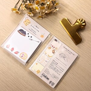 May include: Two packs of sticky memo pads with food illustrations, including strawberries, blueberries, and bread. A gold-coloured paper clip and a bouquet of yellow flowers are also in the image.
