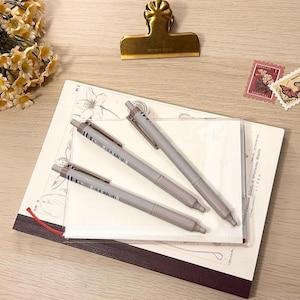 Puede incluir: Vista aérea de artículos de papelería sobre una superficie de madera clara. Tres bolígrafos grises están encima de un cuaderno y una funda de plástico transparente. También hay un clip dorado, dos sellos y un ramo de flores amarillas.