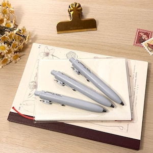 May include: Three pale grey pens lie on a stack of notebooks and stationery. A gold-coloured clip and two decorative stamps are also present. A bouquet of yellow and white flowers is in the upper left corner. The items are arranged on a light wood surface.