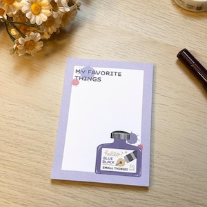 Puede incluir: Un bloc de notas lila con el texto "MY FAVORITE THINGS" y una ilustración de un tintero. El tintero dice "hello? BLUE BLACK SMALL THINGS!" y tiene una capacidad de 50 ml. Artículos de papelería rodean el bloc de notas.