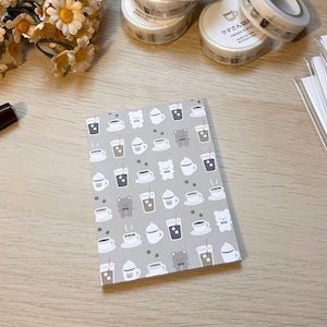 Puede incluir: Un cuaderno gris con un patrón repetido de tazas de café, cafés con hielo y osos de peluche. El cuaderno está rodeado de rollos de cinta adhesiva decorativa y un pequeño ramo de flores. El cuaderno es un artículo de papelería.