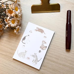 Puede incluir: Una tarjeta blanca con ilustraciones de gatos, tazas de café y un clip dorado. También hay un bolígrafo marrón oscuro y un ramo de flores blancas y amarillas. Las ilustraciones de gatos están en varias poses.