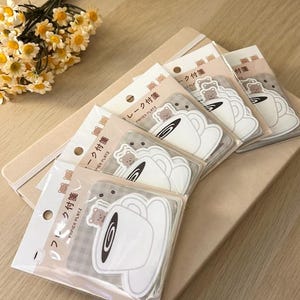 Puede incluir: Varios paquetes de pegatinas con un diseño de taza de té blanca y un oso. Las pegatinas están en envases de plástico transparente, apiladas sobre un cuaderno beige. Un ramo de flores amarillas y blancas está en el fondo.