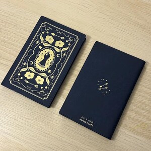 Peut inclure: Deux carnets bleu marine. L'un présente un motif floral et céleste doré avec une figure centrale. L'autre a un motif de cuillère et d'étoile doré. Le texte "PAPIER PLATZ" est visible sur le second carnet.