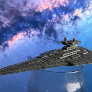 May include: A dark grey model of a Star Destroyer spacecraft, resting on a clear glass jar. The background features a vibrant, colourful nebula with shades of blue, purple, and pink, creating a space-themed aesthetic.