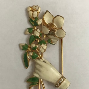 May include: A gold-tone brooch featuring a hand holding a bouquet of white flowers with green leaves. The flowers are enameled in white and the leaves are enameled in green. The hand is also enameled in white and has a row of clear rhinestones around the wrist.