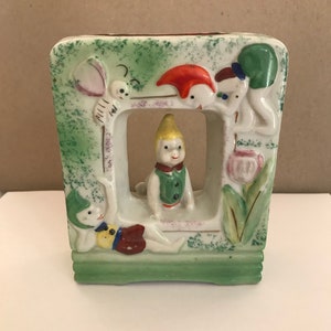 May include: A green ceramic planter with a square shape and a window in the front. The planter features a scene of four whimsical figures with colorful clothing and a butterfly.