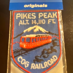 May include: A blue patch with the text "PIKES PEAK ALT. 14,110 FT." and "COG RAILROAD." The patch features a red cog railway train and a mountain scene. The patch is in a clear plastic sleeve with the "Voyager originals" logo.