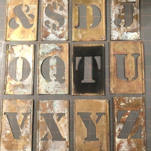 May include: A set of 12 brass stencils featuring the letters & $, D, J, O, Q, T, U, V, X, Y, and Z. The stencils are worn and have a patina finish.