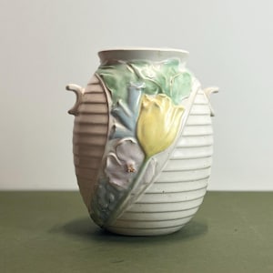 May include: Cream-colored ceramic vase with a ribbed design and floral relief. The vase features a yellow tulip, green leaves, and other pastel-colored flowers. It has two small handles on either side.