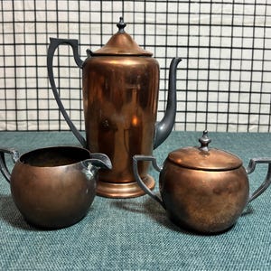 May include: A vintage copper tea set with a teapot, creamer, and sugar bowl. The set has black handles and a shiny copper finish.