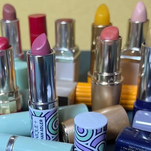 May include: An assortment of lipsticks in various shades, including pink, red, and gold. The lipsticks have metallic and patterned tubes. One lipstick tube has the text "NIQUE+ HAN ADLER". The background is a mix of colors.