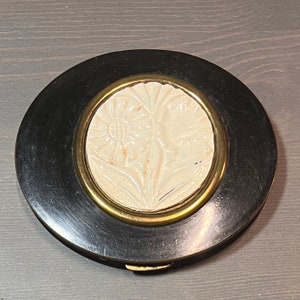 May include: A vintage black and gold compact with a white floral design. The compact has a hinged lid and a small mirror inside.