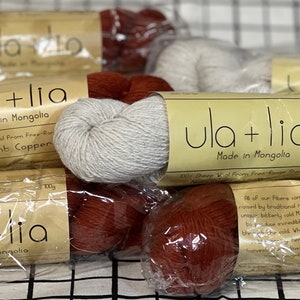 May include: Four skeins of yarn in brown and white colors. The yarn is packaged in clear plastic bags with labels that read "ula+lia Made in Mongolia". The labels also include information about the type of wool used, such as "100% Baby Camel Wool From Free-Range" and "100% Sheep Wool From Free-Range Sheep".