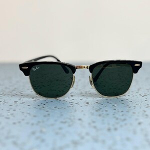Op de afbeelding: Zwarte en goudkleurige zonnebril met donkergroene glazen. De zonnebril heeft een klassiek ontwerp met een zwart bovenframe en goudkleurige metalen accenten. Het Ray-Ban logo is zichtbaar op de linker lens.