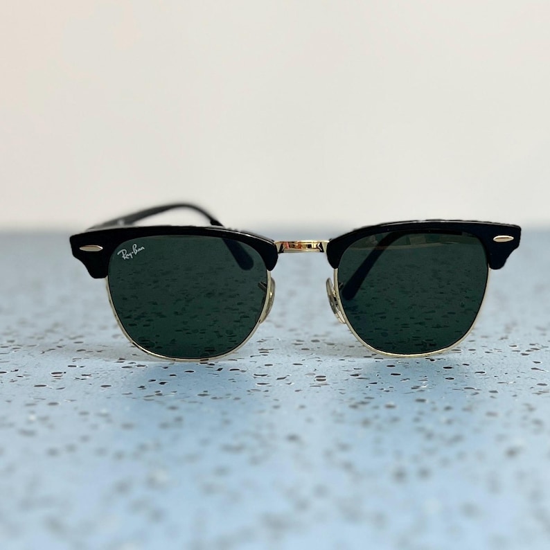 Op de afbeelding: Zwarte en goudkleurige zonnebril met donkergroene glazen. De zonnebril heeft een klassiek ontwerp met een zwart bovenframe en goudkleurige metalen accenten. Het Ray-Ban logo is zichtbaar op de linker lens.